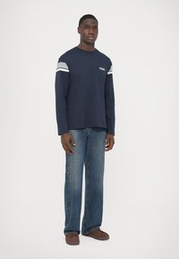 Navy long-sleeve shirt with grey and white stripes on shoulders, paired with light blue straight-leg jeans and brown shoes. Plain background.