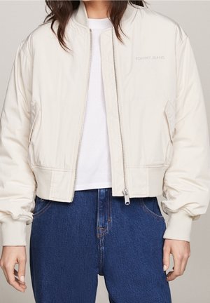 Mujer con una chaqueta bomber blanca de Tommy Jeans sobre una camiseta blanca y unos vaqueros azules de cintura alta, de pie con los brazos relajados.