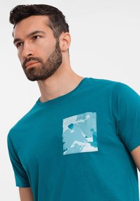 Teal cotton t-shirt featuring a large, blue camo-patterned pocket on the left side. Short sleeves and a crew neckline. Smooth texture.