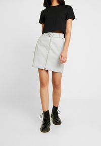 Grijze neplederen rok met een ritssluiting aan de voorkant en zilveren hardware, gecombineerd met een zwart cropped t-shirt en zwarte veterschoenen tot de enkels.
