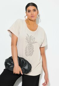 Beige krátký rukáv tričko s grafickým motivem ananasu z kamínků na přední straně, v kombinaci s měkkou černou příruční taškou a černými kalhotami.