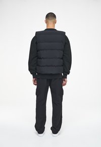 Black puffer vest with horizontal quilting, worn over a black long-sleeve shirt, paired with loose black cargo pants and white sneakers.