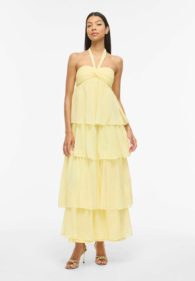 Femme portant une robe maxi à étages jaune pâle avec dos nu croisé et corsage torsadé, associée à des sandales à talons hauts dorées à brides, se tenant devant un fond blanc.