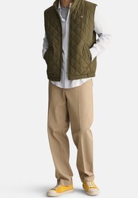 Quilted olive green vest over a gray shirt, paired with tan pants and yellow sneakers. Features a zip closure and side pockets.