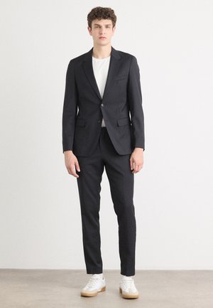 Black tailored suit with notch lapels, single-button closure, and two front pockets. Worn over a white shirt, paired with white sneakers.