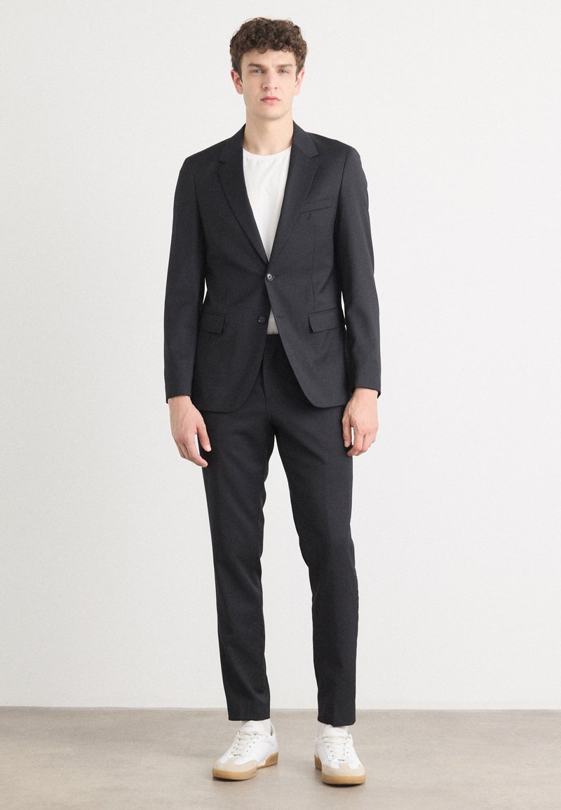 Mustriline mustükk, millel on sisselõigatud krae, ühe nupuga kinnitamine ja kaks eesmist taskut. Kantud valge särk koos valgete tossudega.