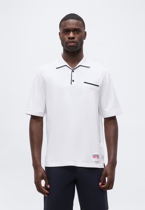Mand iført hvid polo med korte ærmer med sort kant og navy shorts, stående mod en ensfarvet hvid baggrund.