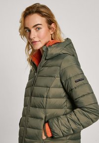 Olijfgroene pufferjack met horizontale quilting, een capuchon en contrasterende oranje voering. Bevat zwart logo op de mouw.