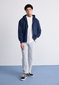 Námornícky modrý zip-up mikina cez biely tričko, svetlo sivé rovné džínsy a čierno-biele tenisky. Hladká textília, voľný strih.