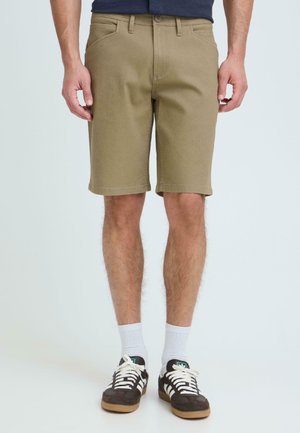 Hombre vistiendo pantalones cortos caqui, zapatillas negras y blancas, y calcetines blancos hasta la altura del tobillo, de pie contra un fondo claro y liso.