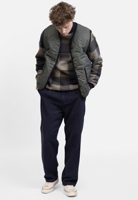 Padded green vest over a patterned sweater, navy trousers, and light sneakers. The vest has a quilted design and side pockets.
