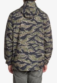 Kurtka wiatrówka z kapturem w kamuflażowym wzorze obejmującym odcienie zieleni i czerni, z elastycznymi mankietami i luźnym krojem. Teksturowane wykończenie materiału.