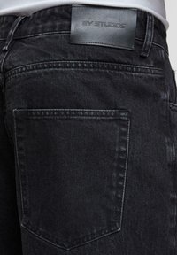 Svart denim jeans har en jevn tekstur, baklomme og en lærmerkel med "AY STUDIOS" brodert. En enkel, funksjonell design.