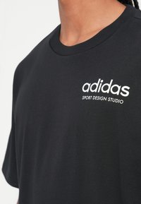 Črna bombažna majica s krožnim izrezom in kratkimi rokavi. Ponaša se z belim logotipom "adidas SPORT DESIGN STUDIO" na levem prsnem predelu.