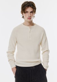 Pulover henley bej, cu mânecă lungă, cu guler rotund, închidere cu trei butoane și croială relaxată. Acesta este asortat cu pantaloni închisi la culoare.