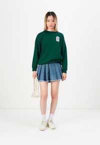 Sweatshirt vert avec un logo 'G' rose, associé à une mini-jupe en jean plissée. Baskets blanches, chaussettes blanches et un petit sac à main crème.