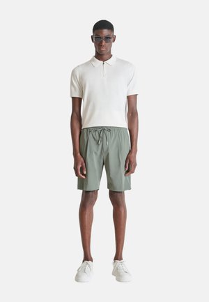 Hombre con gafas de sol oscuras, polo blanco de manga corta con cremallera, pantalones cortos verdes con cordón y zapatillas blancas, de pie mirando hacia adelante.