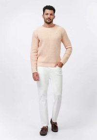 Ποικιλόχρωμο πουλόβερ σε ροδακινί αποχρώσεις με ριμπ μανσέτες και τελείωμα, συνδυασμένο με λευκό παντελόνι και καφέ παπούτσια. Το πουλόβερ έχει ένα μικρό λογότυπο στο στήθος.