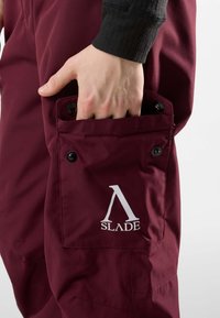 Pantaloni da lavoro bordeaux con tasca laterale, bottoni neri e logo bianco. Il tessuto appare liscio e resistente. Mano inserita nella tasca.