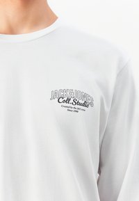 Balta ilgomis rankovėmis marškiniai su užrašu "JACK&JONES Coll. Studio Sukurtas J&J komandos nuo 1990" spausdintu ant kairiojo krūtinės.