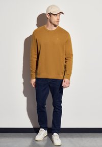 Senfgelbes Sweatshirt mit Rundhalsausschnitt, kombiniert mit dunklen Jeans und weißen Sneakers. Modell trägt eine hellbeige Mütze. Einfaches Design, keine Muster.