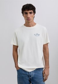 Cremefarbenes Baumwoll-T-Shirt mit einem blauen Logo auf der Brust. Gerade geschnittener Stil mit kurzen Ärmeln, kombiniert mit ausgewaschenen blauen Jeans.