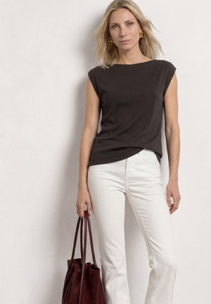 Femme aux cheveux blonds portant un haut noir sans manches et un pantalon blanc, tenant un grand sac à main marron foncé, debout contre un mur blanc.