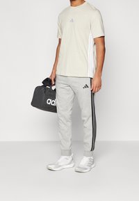 Světle šedé sportovní tričko s malým logem, spárované se šedými tepláky se černými pruhy a taškou Adidas. Bílé tenisky.