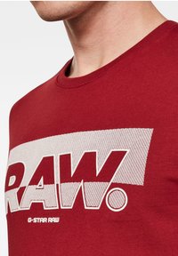 Red cotton t-shirt featuring a large white "RAW." graphic with a textured background. Crew neck design without any additional embellishments.