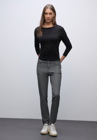 Graue, taillierte Hose mit vorderen Taschen, Knopfverschluss und Nähdetails. Kombiniert mit einem schwarzen Langarmoberteil und weißen Sneakers.
