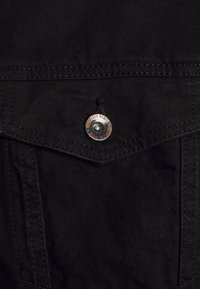 Tissu en denim noir avec une poche cousue. Présence d'un bouton en métal gravé "Refined Rebel". Texture lisse avec des détails subtils.