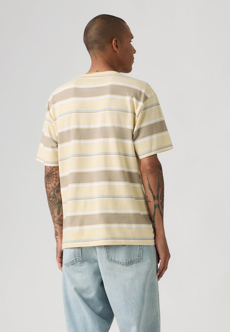 Homme tatoué portant une chemise à manches courtes rayée beige et crème et un jean bleu clair, vu de dos sur un fond uni.
