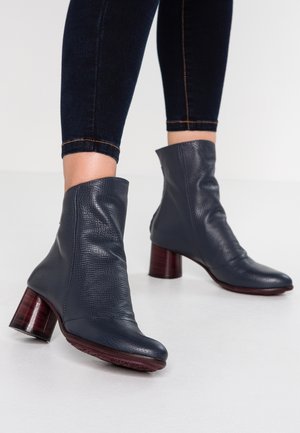 Femme portant des bottines en cuir texturé foncé à talons carrés, associées à un pantalon cintré foncé, sur un fond uni.