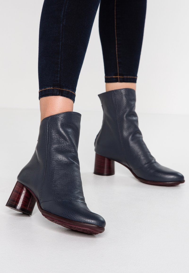 Femme portant des bottines en cuir texturé foncé à talons carrés, associées à un pantalon cintré foncé, sur un fond uni.
