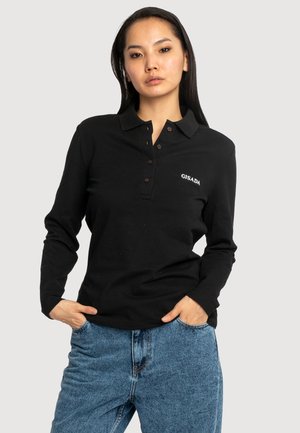 Femme aux longs cheveux foncés portant un polo noir à manches longues avec le texte "GISADA" et un jean bleu, debout les mains dans les poches.