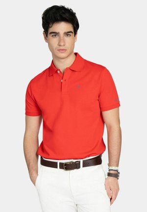 Joven con polo rojo de manga corta metido en pantalones blancos con cinturón marrón, posando frente a un fondo liso.