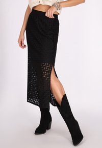 Schwarzer, gestrickter Maxirock mit geometrischem Muster, ausgestattet mit einem seitlichen Schlitz und einer strukturierten Oberfläche, kombiniert mit schwarzen Wildleder-Stiefeletten.