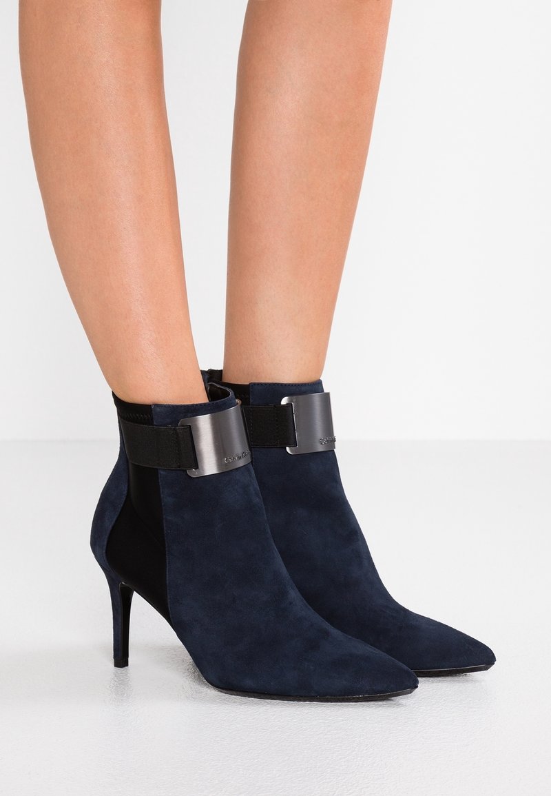 Femme portant des bottines à talons hauts en daim bleu marine, à bouts pointus et avec un détail de sangle métallique autour des chevilles.