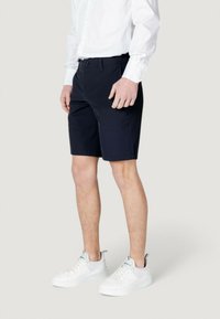 Pantaloni corti blu navy con una texture liscia e una vestibilità su misura, dotati di tasche laterali e chiusura a bottone frontale, abbinati a sneakers bianche.