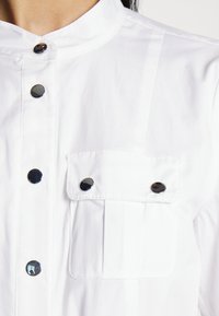 Camicia bianca con colletto alla coreana, bottoni a pressione neri lucidi e taschino sul petto con patta, che mette in risalto una superficie di tessuto liscia.