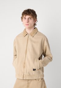 Beige denimpintsak, millel on ees lukk, langetatud krae, küljetaskud ja väike logopatch ääres. Sile tekstuur.