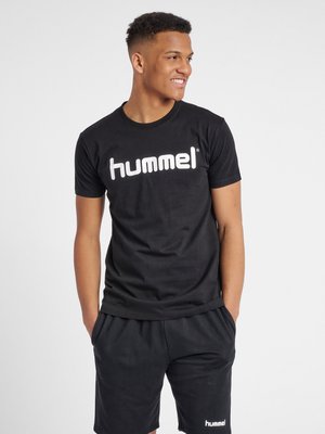 Young man smiling, hands in pockets, wearing black Hummel t-shirt and shorts against plain light background.