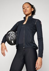 Black cycling jacket with a zippered front, long sleeves, and textured shoulder panels. Model holds a black helmet. Plain background.