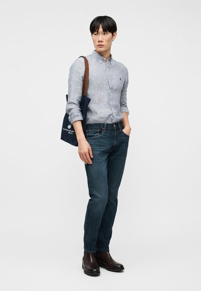 Νεαρός άνδρας που φοράει ριγέ πουκάμισο με κουμπιά, μπλε τζιν, καφέ μπότες, κρατώντας ένα σκούρο μπλε τσάντα χειρός με καφέ λουριά, στέκεται μπροστά σε απλό φόντο.