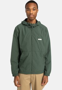 Grüne leichte Jacke mit einem Reißverschluss vorne, verstellbarem Kapuzen und elastischen Bündchen. Hat ein kleines Logo und Seitentaschen.