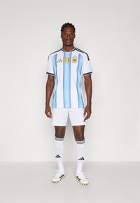 Zila un baltā svītraina futbola jersey ar melnām akcentiem, zelta emblemas, baltas bikses, melnas un baltas svītraines zeķes, un melni sporta apavi.