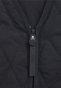 Jachetă neagră matlasată cu un fermoar central, dotată cu un tab de prindere în formă de craniu și oase încrucișate, material texturat și cusături în model de diamant.