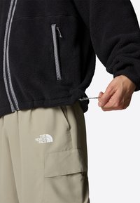 Svart fleecejacka med en zip-pocket, grå accenter och justerbar dragsko i fållen; ihopsatt med beige cargobyxor med en logotyp.