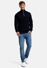 Marineblauer Strickpullover mit hohem Kragen und einem Reißverschluss auf der Vorderseite, kombiniert mit hellblauen Jeans und weißen Sneakers, die ein strukturiertes Design aufweisen.