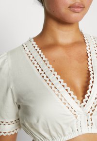 Γυναίκα που φοράει ένα άσπρο μπλουζάκι με ζιγκ-ζαγκ δαντελένια επένδυση κατά μήκος του βαθύ V-λαιμού και κοντά μανίκια.